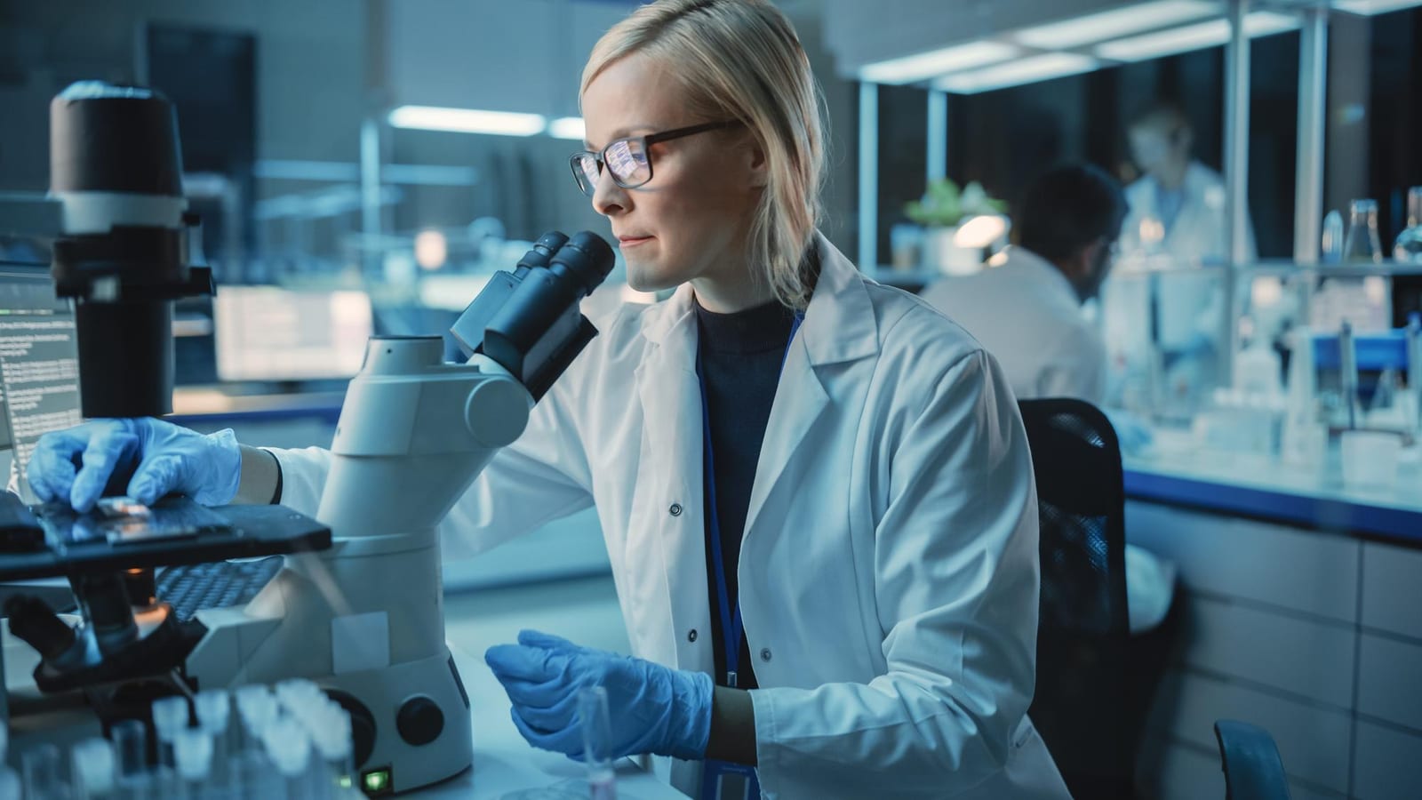 Pesquisadora de jaleco branco observando amostra em microscópio sob luz azulada